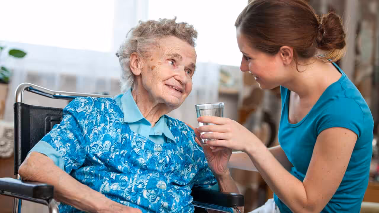 A smiling elderly woman in a wheelchair receives a glass of water from a younger woman who is crouched beside her, both making eye contact in a warm, caring interaction.