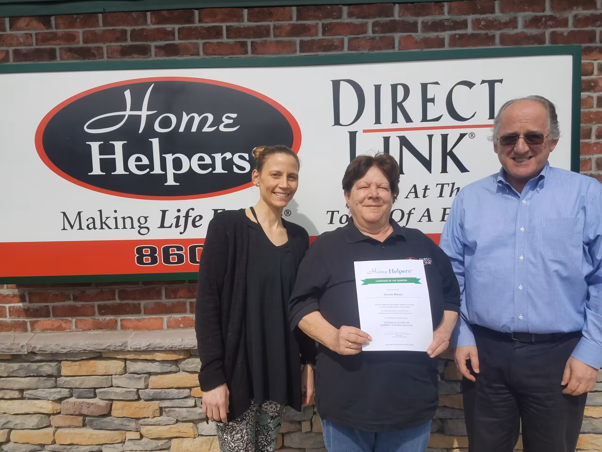 Three people stand in front of a Home Helpers and Direct Link sign. The person in the center is holding a certificate and smiling, while the other two stand on either side, also smiling. The background is a brick wall.