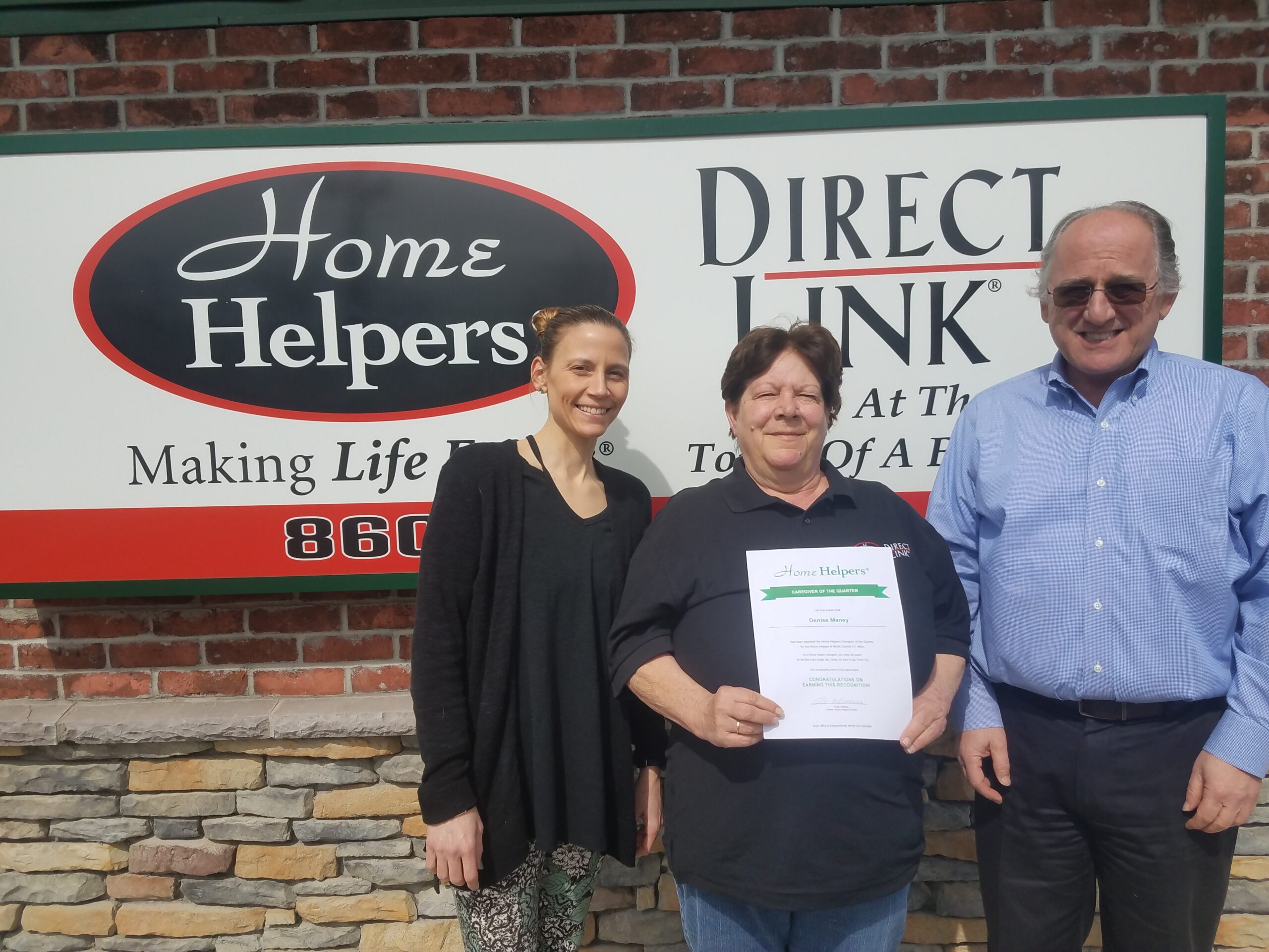 Three people stand in front of a Home Helpers and Direct Link sign. The person in the center is holding a certificate and smiling, while the other two stand on either side, also smiling. The background is a brick wall.