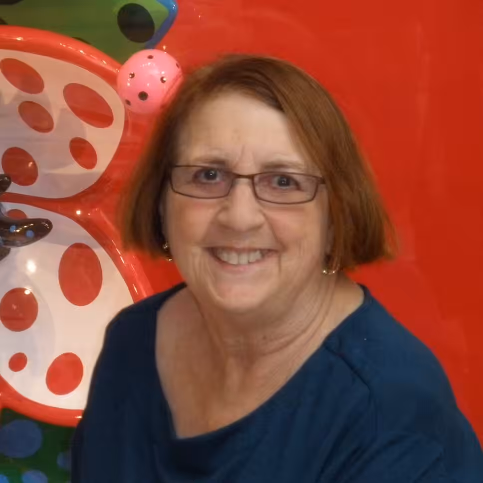 A smiling older woman with short brown hair and glasses in front of a bright red background with a large, colorful ladybug decoration.