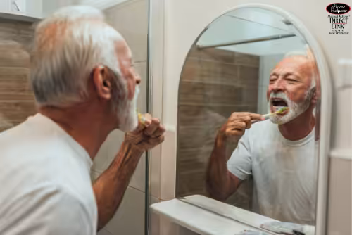 An older man with gray hair and a beard is standing in front of a bathroom mirror, brushing his teeth while wearing a white t-shirt. The background shows tiled walls and a sink.