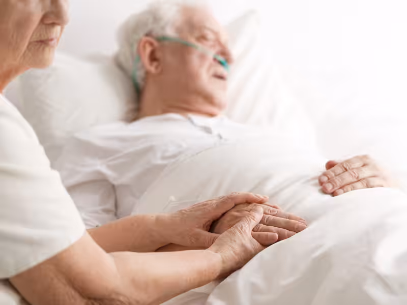 An elderly person lies in a hospital bed with an oxygen tube, while another person sits beside them, gently holding their hand in a comforting gesture.