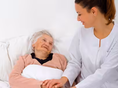 A smiling nurse in a white uniform holds the hand of an elderly woman lying in bed, both looking at each other warmly in a bright, peaceful room.