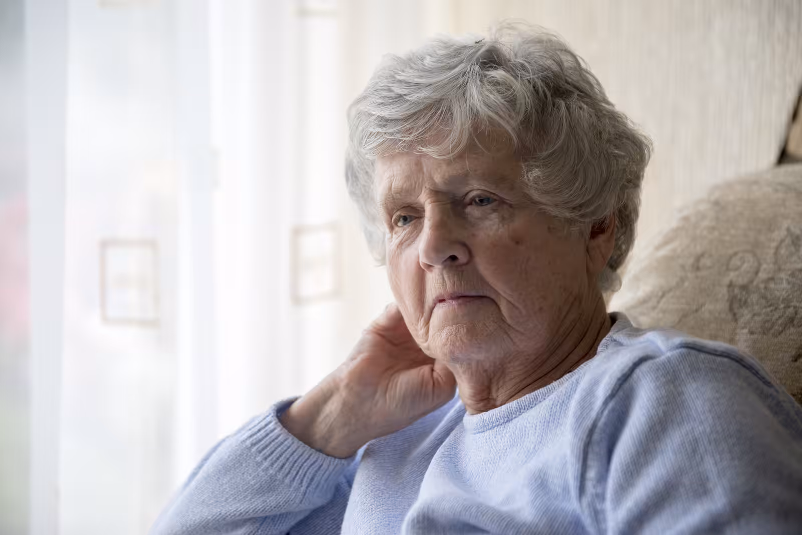 An elderly woman with short gray hair sits indoors by a window, resting her hand on her neck and looking thoughtfully outside. She wears a light blue sweater and appears pensive or contemplative.