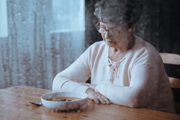 An elderly woman sits alone at a table, looking down at a bowl of soup. Rain is visible on the window beside her, creating a somber mood.