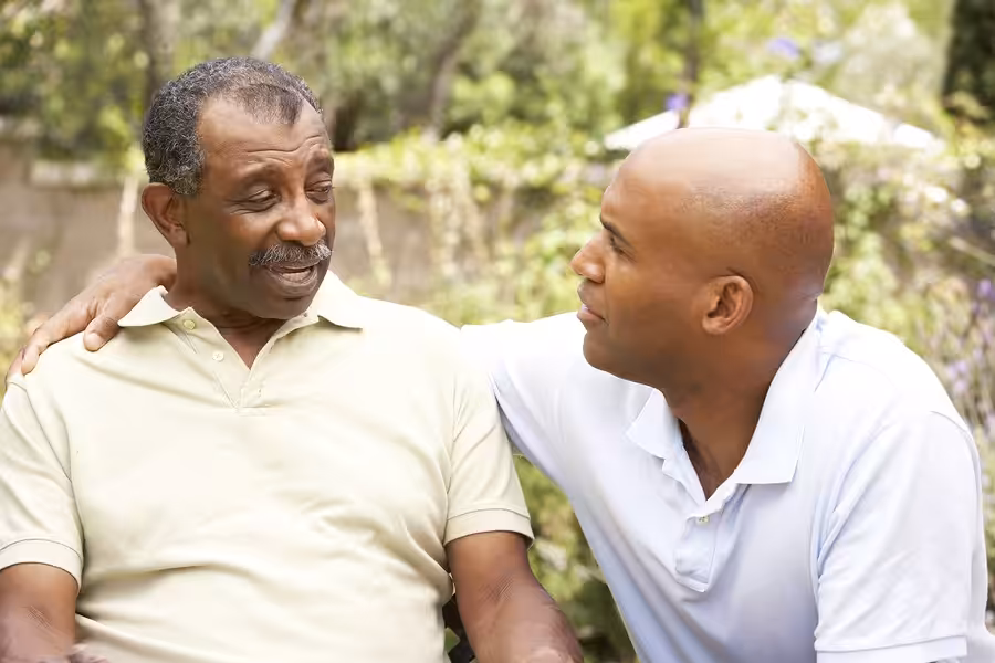 Two men sitting outside, one older and one younger, are having a conversation. The younger man has his arm around the older mans shoulder, and they look at each other with warm, supportive expressions. Trees and greenery are in the background.