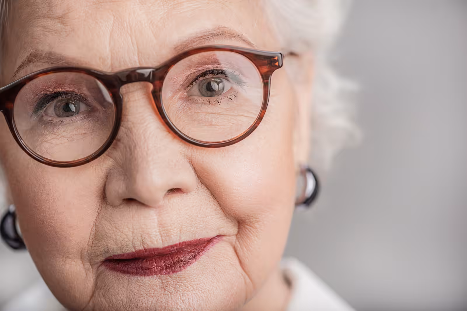 Close-up of an elderly woman with white hair, wearing round eyeglasses and red lipstick, looking directly at the camera with a gentle expression against a neutral background.