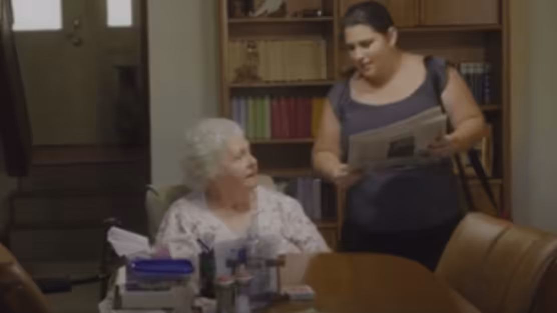A younger woman hands a newspaper to an older woman sitting at a dining table, which holds various household items. Bookshelves and a door are visible in the background.