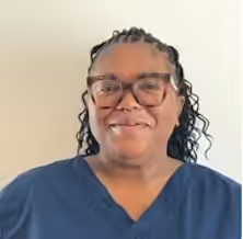 A woman with medium skin tone, wearing glasses and a navy blue scrub top, smiles at the camera in front of a plain light-colored wall. She has long, curly hair styled with small braids.