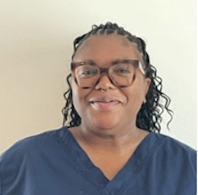 A woman with medium skin tone, wearing glasses and a navy blue scrub top, smiles at the camera in front of a plain light-colored wall. She has long, curly hair styled with small braids.