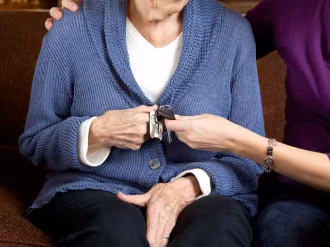 An elderly person in a blue cardigan sits while another person hands them a set of keys. The elderly persons hands rest in their lap, and the other persons arm offers gentle support.