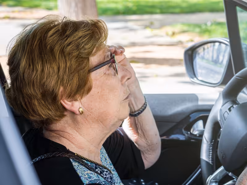 An elderly woman with short hair and glasses sits in the driver’s seat of a car, holding her hand to her forehead and looking ahead, appearing thoughtful or concerned.