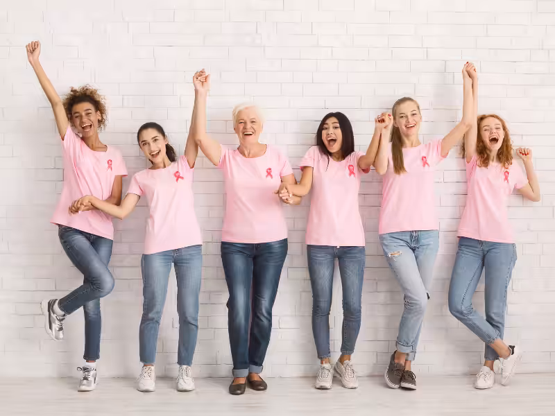 Six women of different ages and ethnicities, wearing pink shirts with pink ribbons and jeans, stand together against a white brick wall, smiling and cheering in support of breast cancer awareness.