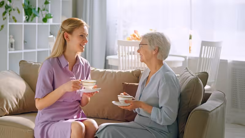 A young woman and an older woman sit on a sofa, smiling and holding teacups. They appear to be enjoying a friendly conversation in a bright, cozy living room.