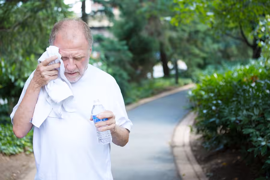 An older man in a white shirt wipes his face with a towel and holds a water bottle while walking on a paved path surrounded by green trees and bushes.
