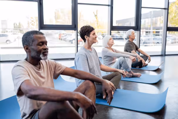 Four adults sit cross-legged on yoga mats in a bright room with large windows, eyes closed and hands resting on their knees, appearing relaxed and peaceful during a group meditation or yoga session.