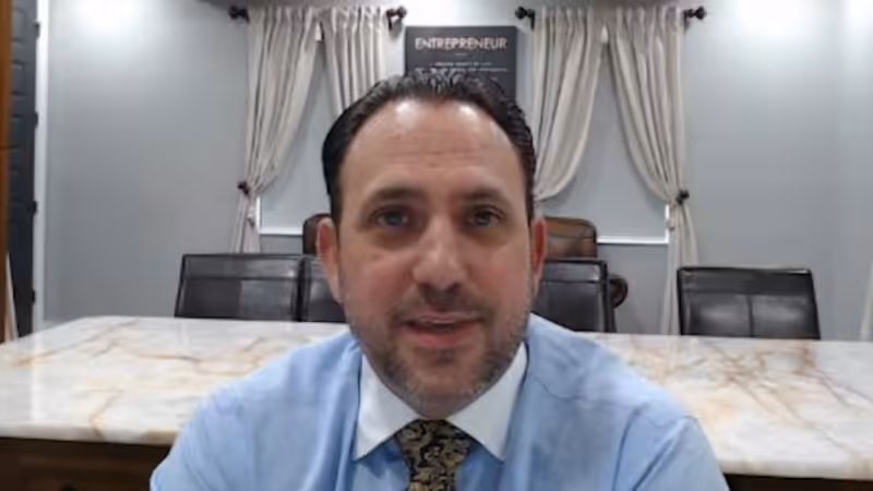 A man with dark hair and a beard, wearing a light blue shirt and patterned tie, sits at a marble conference table with beige curtains and a sign that reads Entrepreneur in the background.