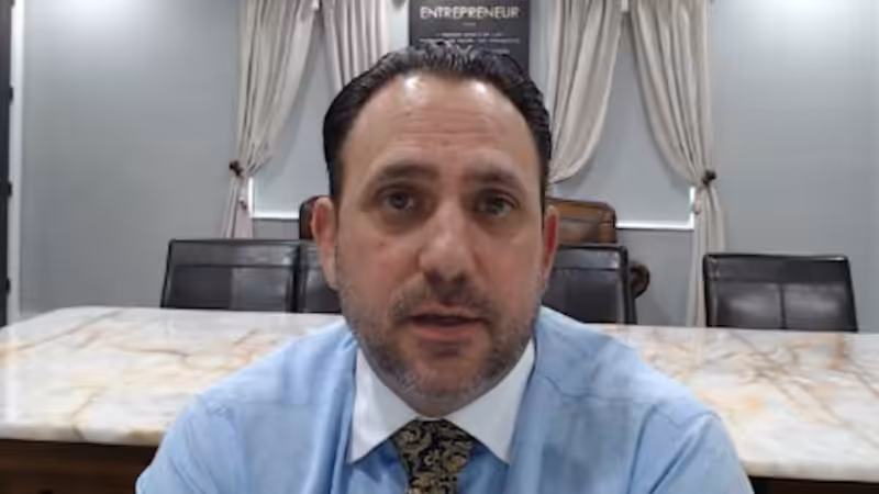 A man with dark hair and a trimmed beard sits at a conference table in an office, wearing a blue shirt and patterned tie. Light curtains and a sign reading ENTREPRENEUR are visible in the background.