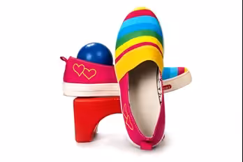 A pair of colorful slip-on shoes with rainbow stripes and pink heels featuring heart designs, displayed with one shoe standing upright and the other resting on a red block and a blue ball, against a white background.