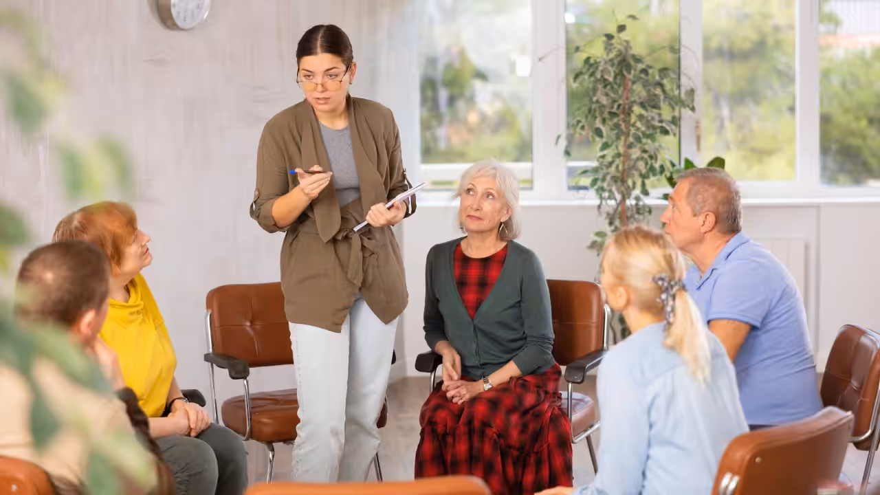 A woman stands in front of a seated group of older adults, holding a notebook and pen while speaking. The group listens attentively in a bright room with large windows and indoor plants.