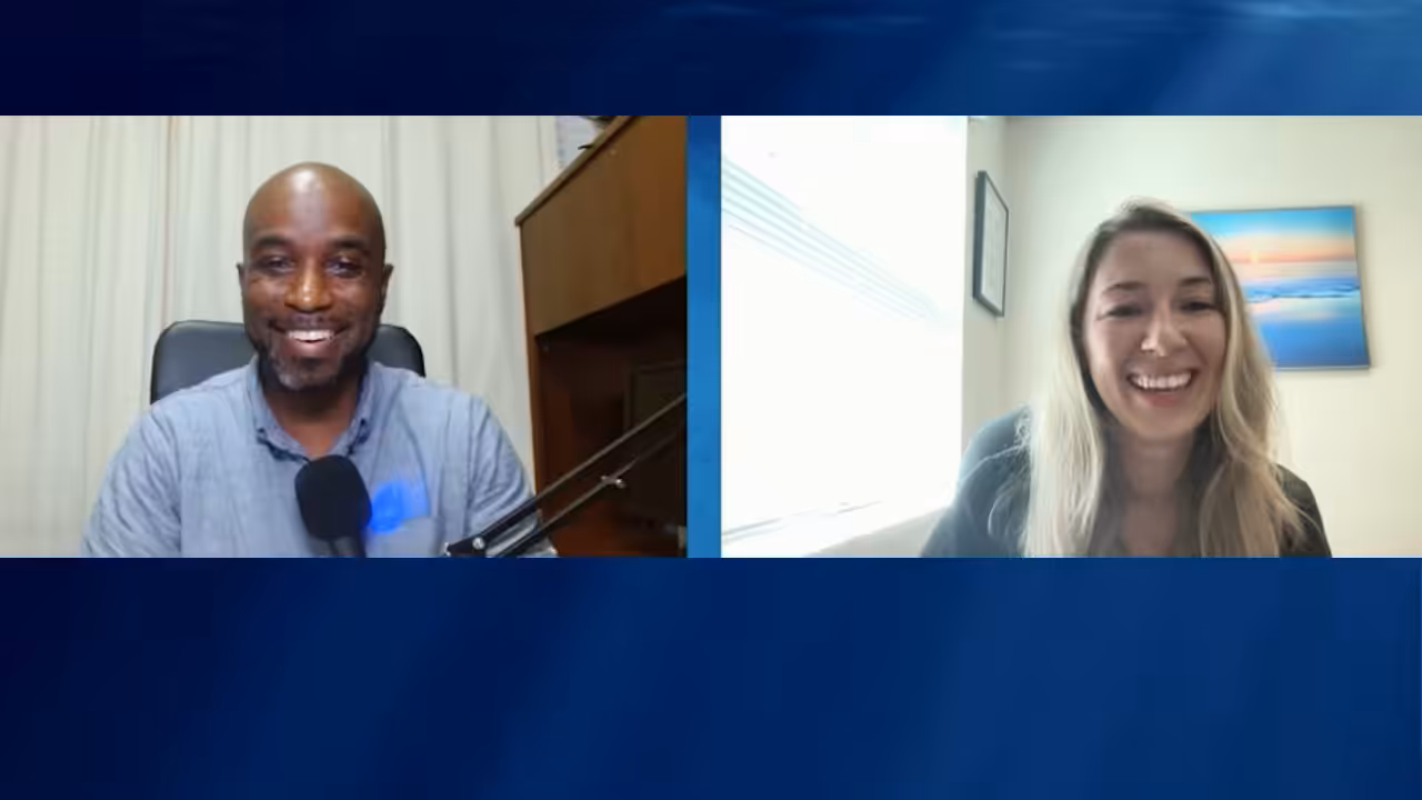 Two people are smiling during a video call. The man on the left has a microphone and sits in front of a curtain and desk, while the woman on the right sits by a window with a framed picture on the wall behind her.