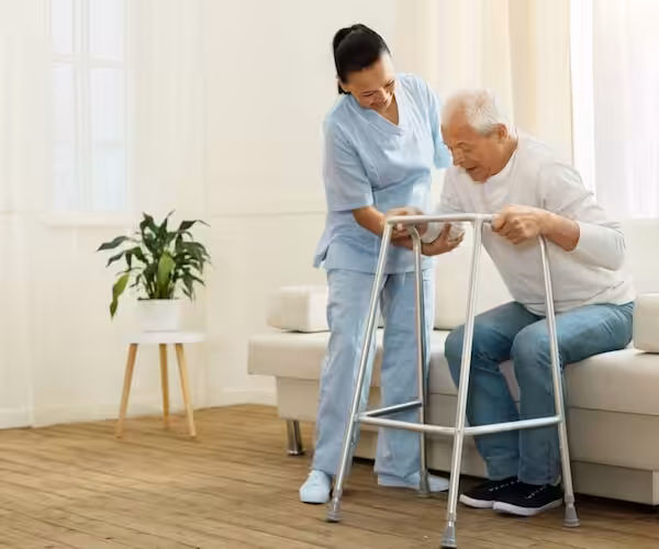 A nurse assists an elderly man using a walker to stand up from a sofa in a bright, cozy living room with a potted plant in the background.