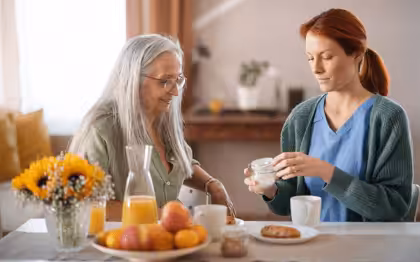 A younger woman helps an older woman at a table with breakfast foods, including juice, fruit, and pastries. The younger woman is holding a jar, possibly medication, while the older woman watches. Sunflowers are on the table.