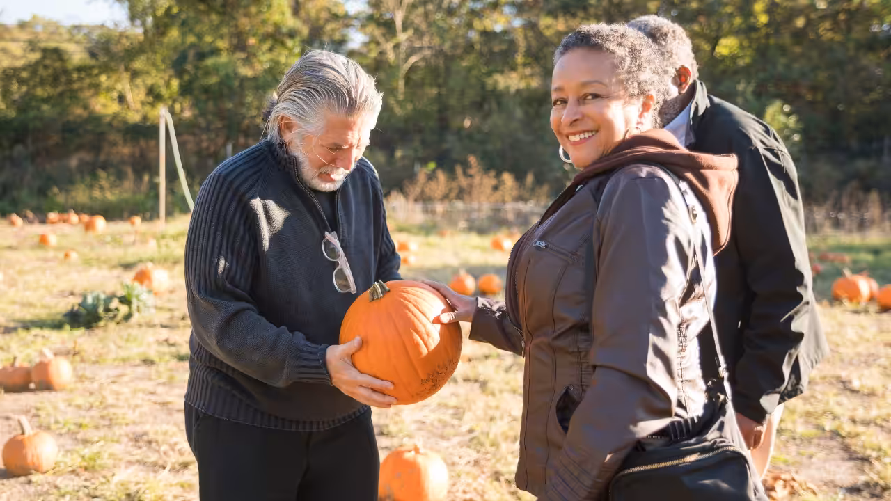 Two adults stand in a pumpkin patch. One man in a dark sweater holds a pumpkin and smiles, while a woman in a brown jacket smiles at the camera. Another person stands behind them. Orange pumpkins are scattered around.