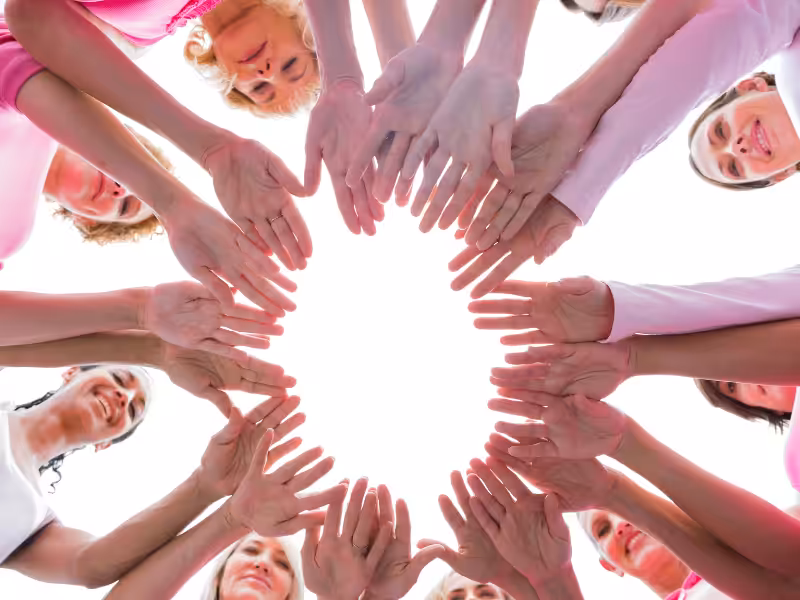 A group of people standing in a circle, reaching their hands toward the center, forming a star shape. They are smiling and wearing pink clothing, symbolizing unity and support.