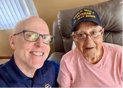 Two older men smile for a selfie indoors. One wears glasses and a dark shirt, while the other wears a cap that reads “World War II Veterans Navy Seabees” and a pink shirt. They are sitting close together on a chair or sofa.