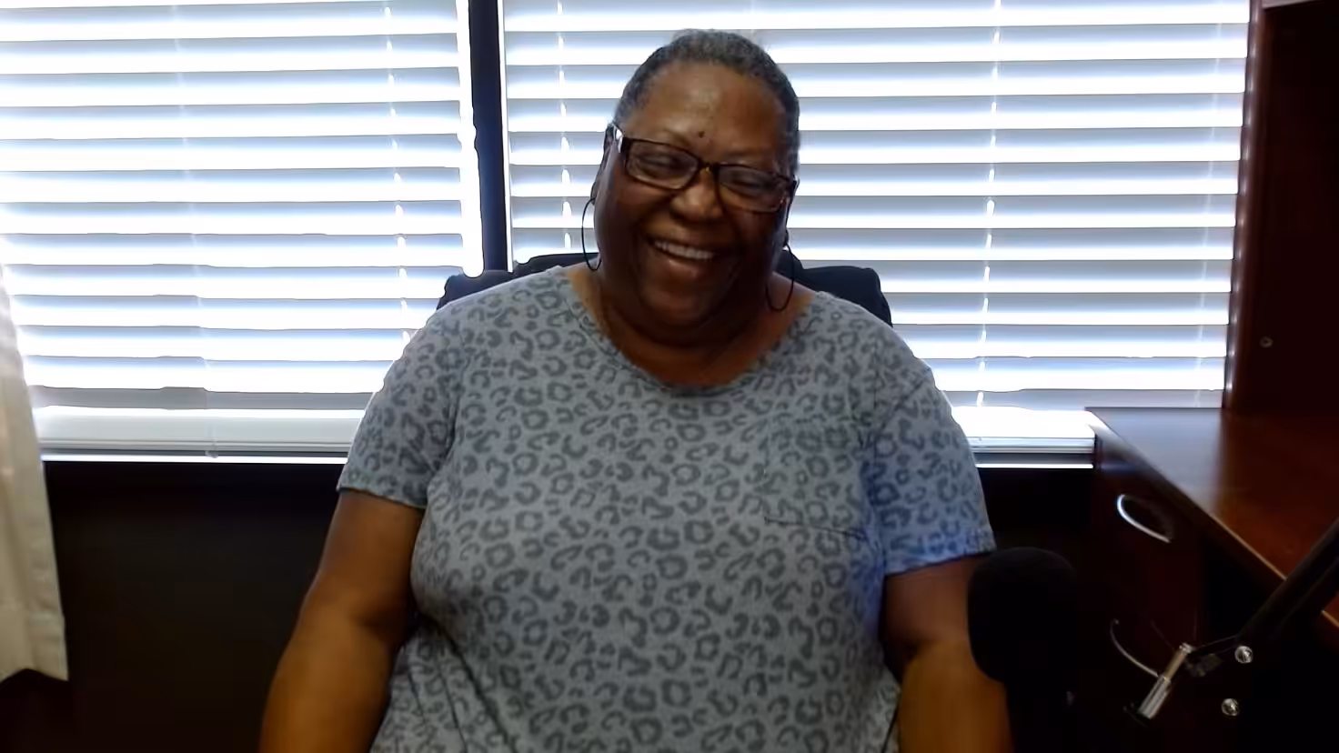 A smiling woman wearing glasses and a gray leopard-print shirt sits in an office chair in front of a window with closed blinds. A microphone is visible in the foreground.
