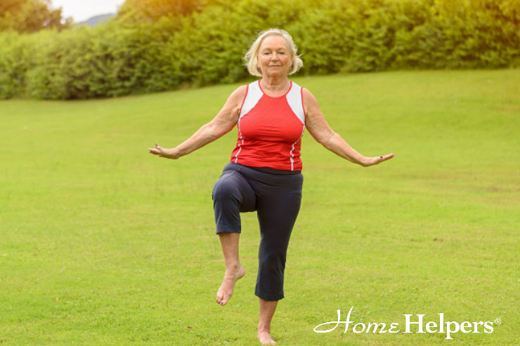 Older woman in athletic wear balances on one leg with arms outstretched in a grassy park, smiling. The Home Helpers logo appears in the bottom right corner.