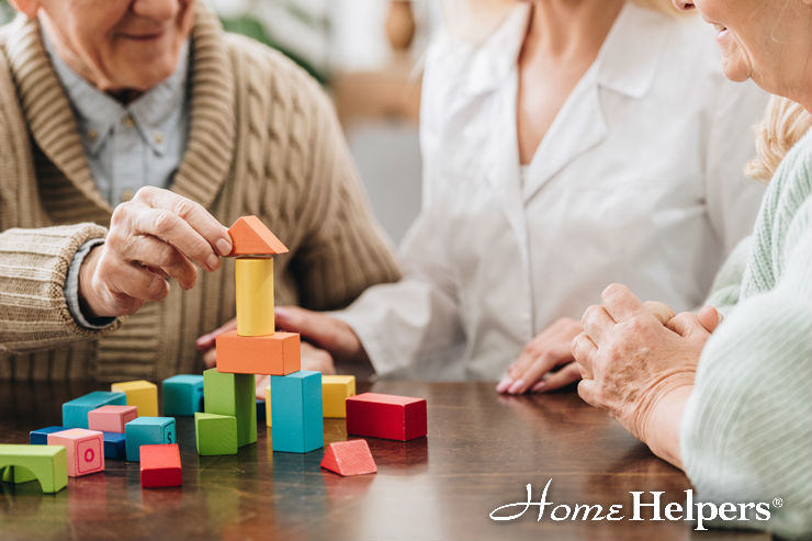 Two seniors and a caregiver sit at a table building a colorful structure with wooden blocks. The seniors appear engaged and smiling, with the caregiver observing. Home Helpers logo is visible in the lower right corner.