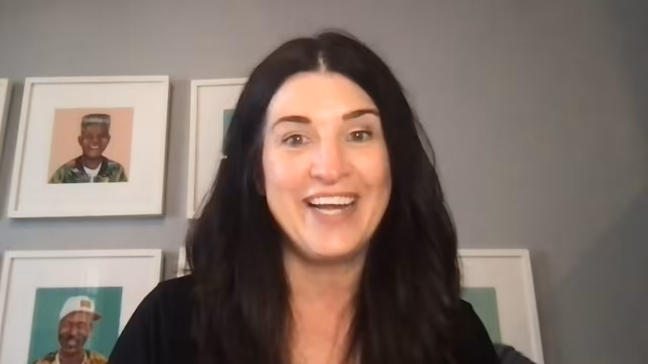 A woman with long dark hair smiles at the camera, with three framed portraits on the wall behind her against a gray background.