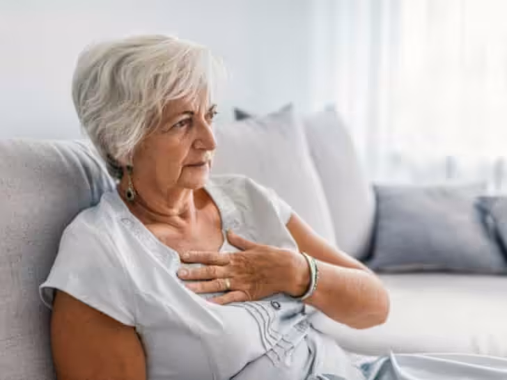 Elderly woman with short white hair sits on a couch, holding her chest with a concerned expression, suggesting possible discomfort or pain. Neutral-toned pillows and soft light create a calm setting.