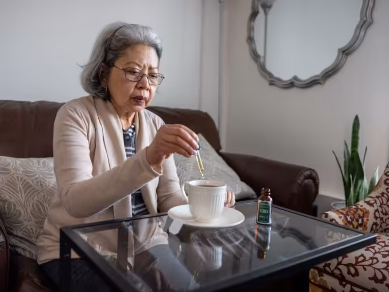 An older woman sits on a couch, adding drops from a CBD oil bottle into a cup on a glass table, with a mirror and plant in the background.