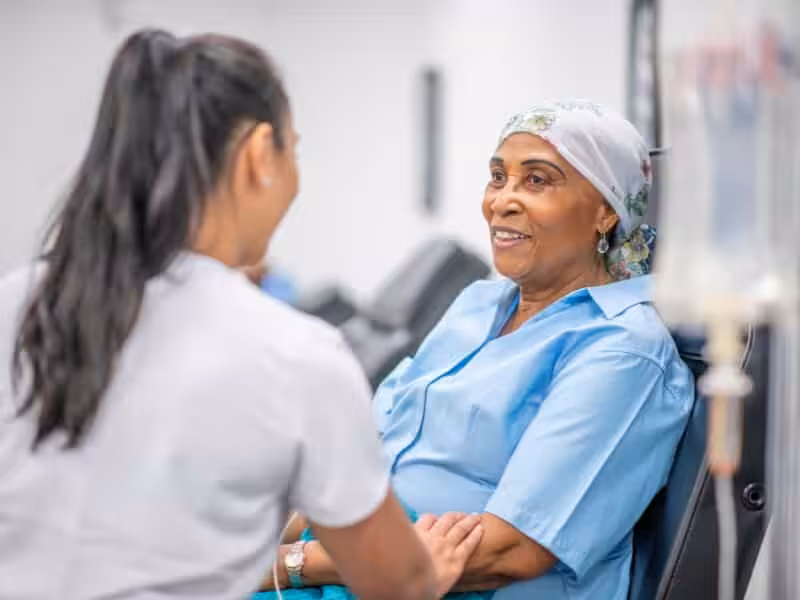 A nurse holds the hand of a smiling older woman wearing a headscarf and blue shirt, sitting in a medical facility, possibly receiving treatment.