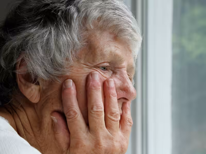 An elderly person with gray hair rests their head on their hand, gazing out of a window with a thoughtful, contemplative expression.