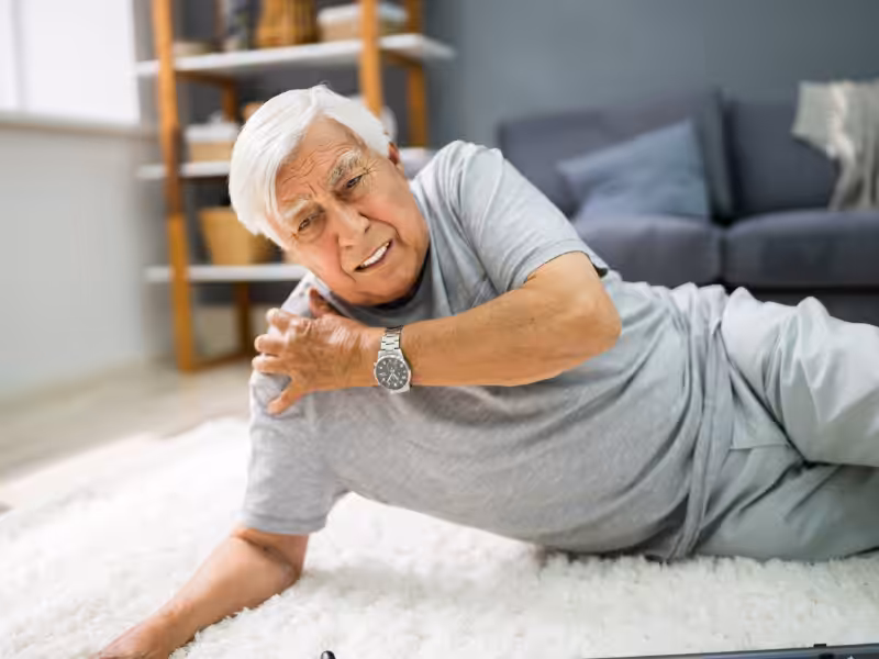 An older man with gray hair lies on a white carpet, grimacing in pain and holding his shoulder, suggesting he has fallen. A sofa and shelves are visible in the background.