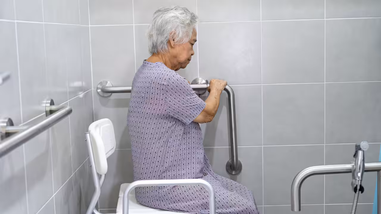 An elderly person with short gray hair sits on a shower chair in a tiled bathroom, holding onto metal grab bars for support. The person wears a patterned hospital gown and faces sideways.