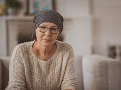 An older woman wearing glasses and a gray headscarf sits on a couch, looking thoughtfully at the camera. She is dressed in a light knit sweater, and the background appears to be a cozy living room.
