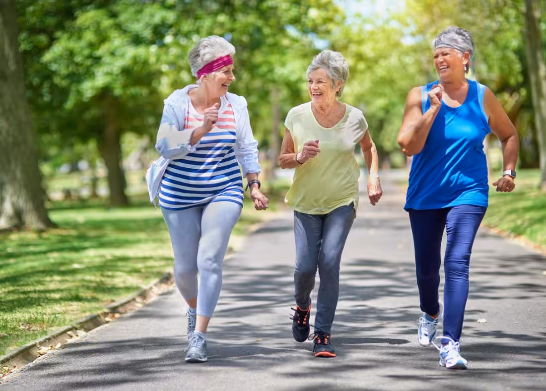 Three older women in athletic clothing are jogging and smiling together on a tree-lined path in a sunny park. The mood is joyful and energetic, with greenery and sunlight in the background.