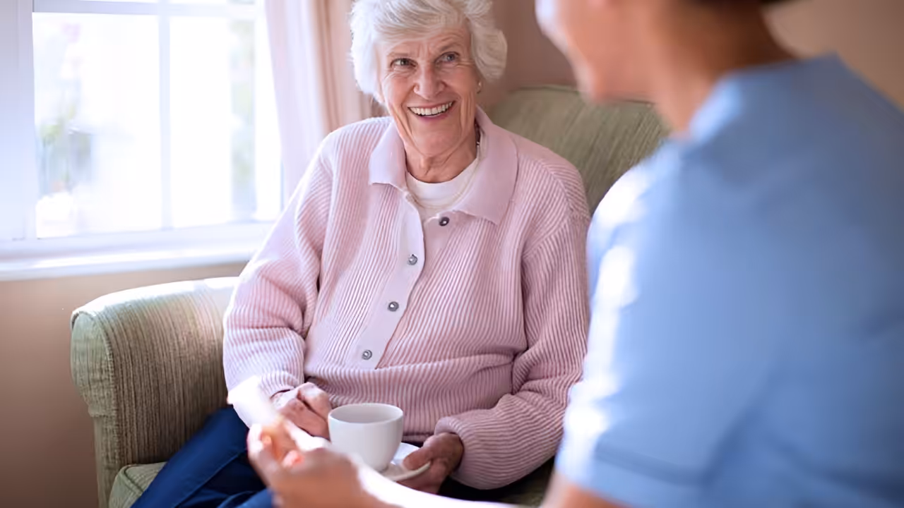 An elderly woman with gray hair smiles while sitting on a sofa, holding a mug. Another person, partially visible in the foreground, appears to be talking to her. Sunlight shines through a nearby window.