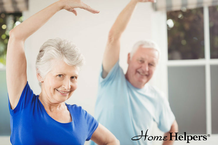 Two smiling older adults in casual clothes are exercising indoors, raising one arm overhead. The woman in front wears a blue shirt; the man behind her wears a light blue shirt. “Home Helpers” logo is in the corner.