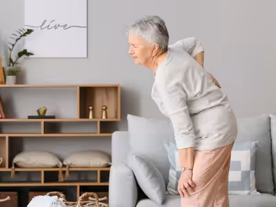 An elderly woman stands beside a sofa, holding her lower back and appearing to be in pain. The room is softly decorated with neutral tones, cushions, and wall art in the background.