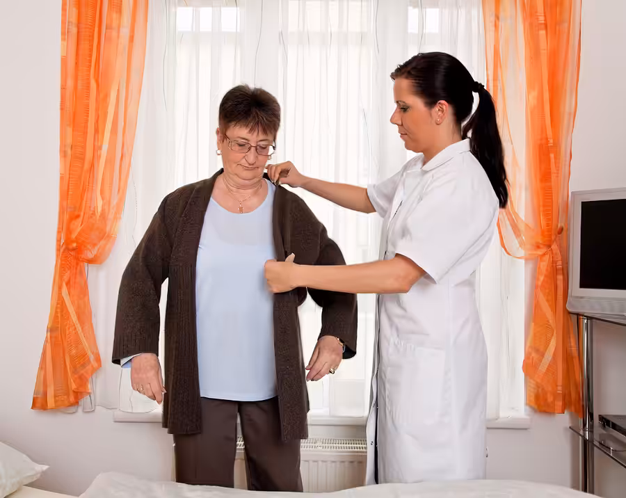 A healthcare worker in a white uniform assists an older woman in putting on a brown cardigan in a bright room with orange curtains and a bed nearby.