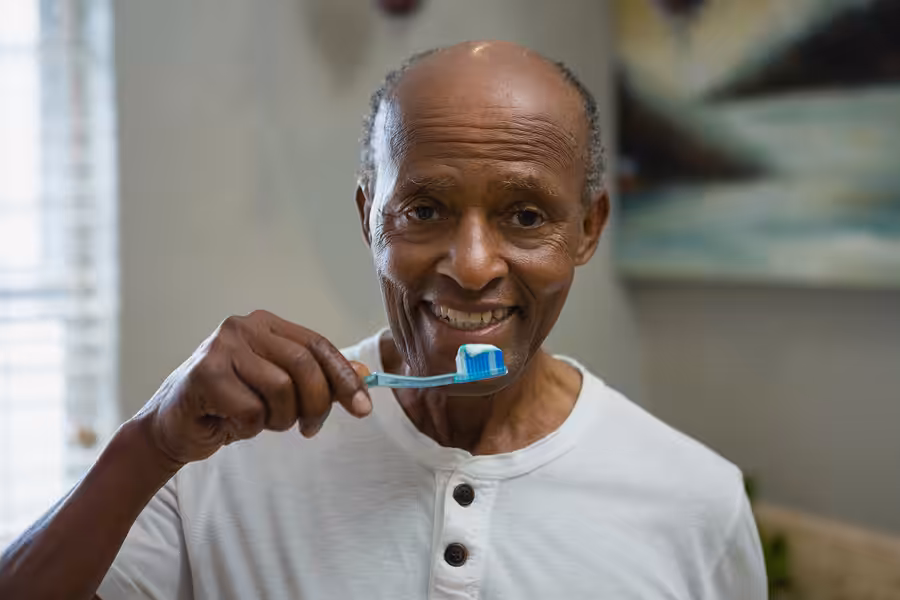 An elderly man in a white shirt smiles while holding a toothbrush with toothpaste, preparing to brush his teeth in a brightly lit room.