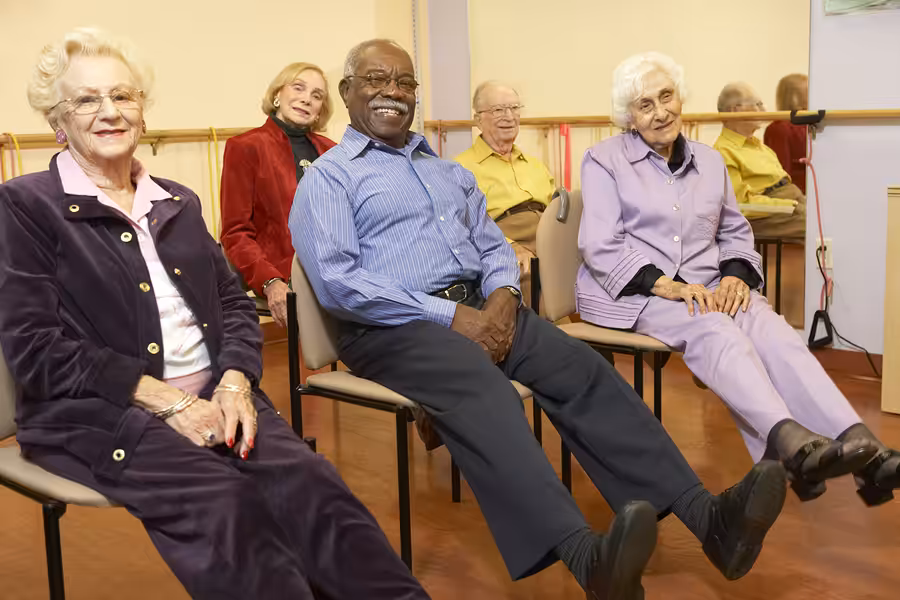A group of five older adults sit on chairs in a row, smiling and lifting their legs in a light exercise class in a bright, indoor setting.