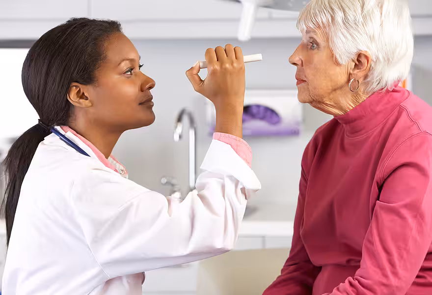 A doctor examines an older woman’s eyes with a penlight in a medical office. The doctor wears a white coat and the patient is dressed in a pink sweater, sitting on the examination table.