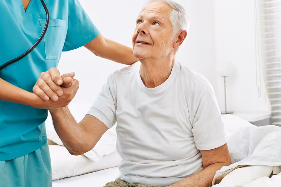 An elderly man sitting on a bed holds hands with a healthcare worker in scrubs, who is offering support. The man looks up at the worker, and the setting appears to be a bright, clean room.